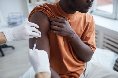 Close up of adult African-American man looking away while getting covid vaccine in clinic or hospital, with male nurse injecting vaccine into shoulder