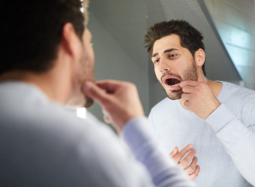 Handsome young man with stubble keeping mouth open while checking tooth and looking into mirror in bathroom