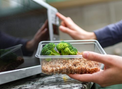 Using the microwave oven to heating food. Woman's hand going to heat up a plastic container with broccoli and buckwheat in the microwave