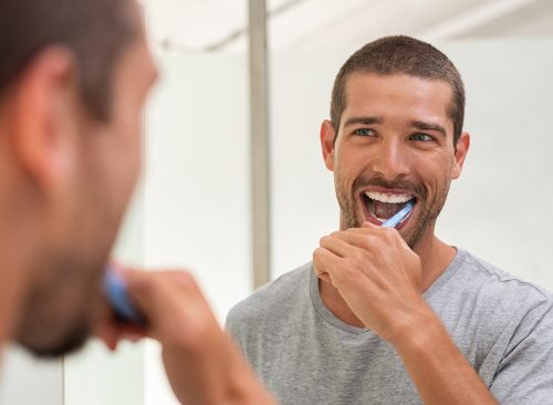 Smiling young man with toothbrush cleaning teeth and looking mirror in the bathroom. Handsome man brushing his teeth in morning in bathroom. Happy guy in pajamas brushing teeth before going to sleep.