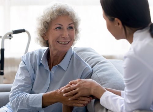 Caring young nurse doctor carer helping holding hands of happy disabled handicapped or injured old adult elder woman having disability health problem sit at home with walking frame walker in hospital