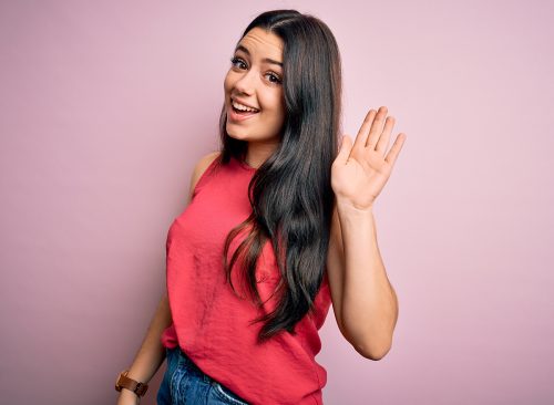 Young brunette woman wearing casual summer shirt over pink isolated background Waiving saying hello happy and smiling, friendly welcome gesture