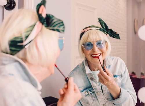 Smiling elderly senior stylish woman in blue sunglasses and denim jacket using red lipstick by the mirror in stylish loft interior. Style, fashion, make up, anti age concept