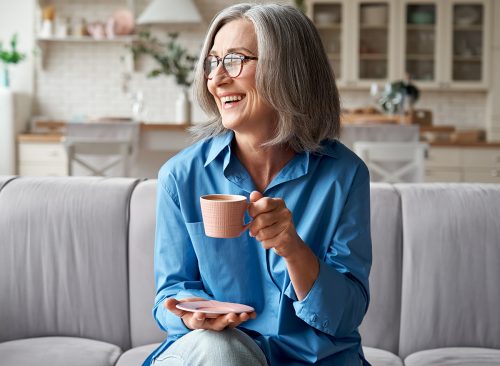 Happy beautiful relaxed mature older adult grey-haired woman drinking coffee relaxing on sofa at home. Smiling stylish middle aged 60s lady enjoying resting sitting on couch in modern living room.