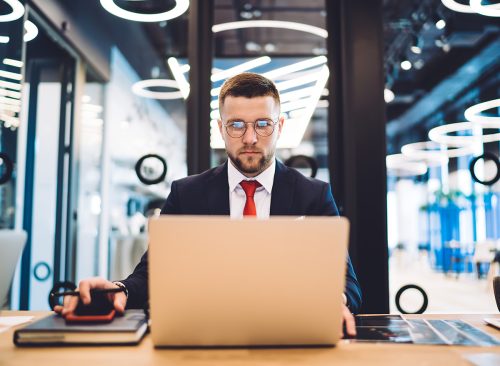 Concentrated bearded male manager in formal wear and eyeglasses sitting at wooden table and working on laptop in modern office