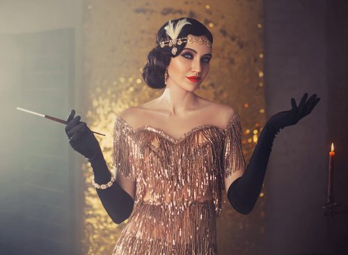 portrait of retro flapper beauty fashion model. Woman holding long slim mouthpiece in hand, cigarette. Party 20s style room full smoke. Gold shiny dress, accessories. Invitation gesture, free space
