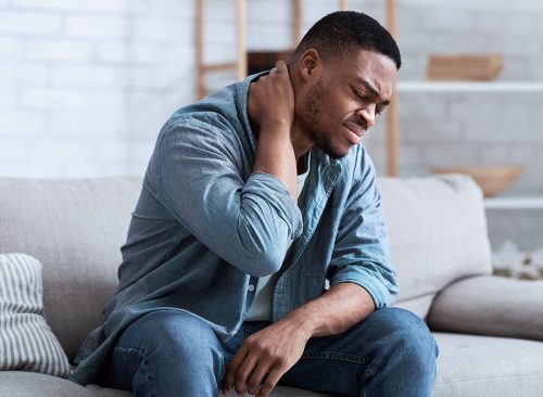 Black Man Suffering From Neck Pain Massaging Aching Muscles Sitting On Couch In Living Room At Home. Neurological Health Problem, Arthritis And Osteoarthritis Concept