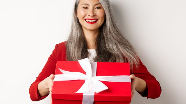 Elegant senior woman giving you present. Asian lady holding red gift box and smiling, standing over white background
