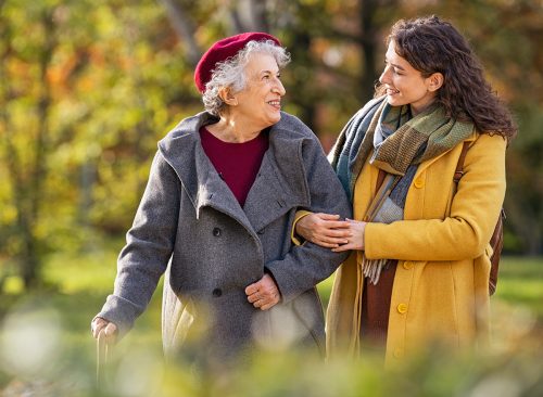 Young woman in park wearing winter clothing walking with old grandmother. Smiling lovely caregiver and senior lady walking in park during autumn and looking at each other with copy space.