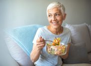 Mature smiling woman eating salad, fruits and vegetables. Attractive mature woman with fresh green salad at home. Senior woman relaxing at home while eating a small green salad, home interior.