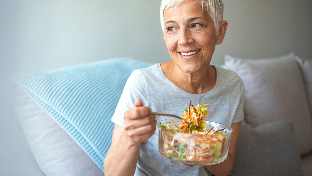 Mature smiling woman eating salad, fruits and vegetables. Attractive mature woman with fresh green salad at home. Senior woman relaxing at home while eating a small green salad, home interior.