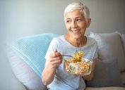 Mature smiling woman eating salad, fruits and vegetables. Attractive mature woman with fresh green salad at home. Senior woman relaxing at home while eating a small green salad, home interior.