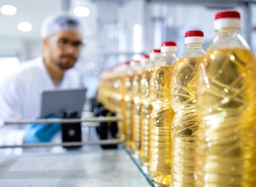 Bottled vegetable oil production in food factory and worker in white coat with hairnet controlling process.