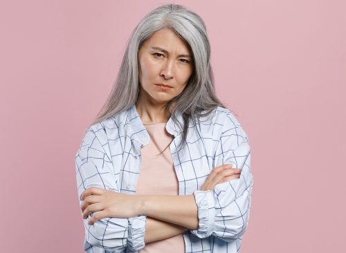 Dissatisfied puzzled concerned gray-haired asian woman wearing basic white checkered shirt standing holding hands crossed looking camera isolated on pastel pink colour background, studio portrait