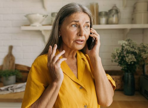 Elderly adorable Caucasian grandmother talking on phone with her grandson standing at kitchen having anxious and worried face expression, looking through window, waiting him for lunch