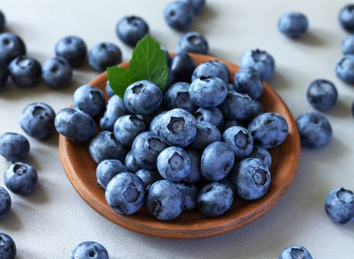 Brown ceramic bowl of fresh ripe blueberries with green leaves on kitchen table