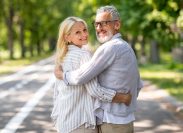 Happy mature spouses embracing while walking in summer park together, smiling senior couple hugging and turning at camera, elderly man and woman enjoying romantic outdoor date, copy space