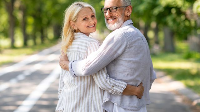 Happy mature spouses embracing while walking in summer park together, smiling senior couple hugging and turning at camera, elderly man and woman enjoying romantic outdoor date, copy space