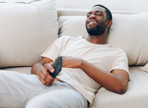 Happy African American Man Relaxing on Black Sofa, Typing Message on Mobile Phone The modern apartment provides a cozy background as he enjoys leisure time, connecting with friends on social media