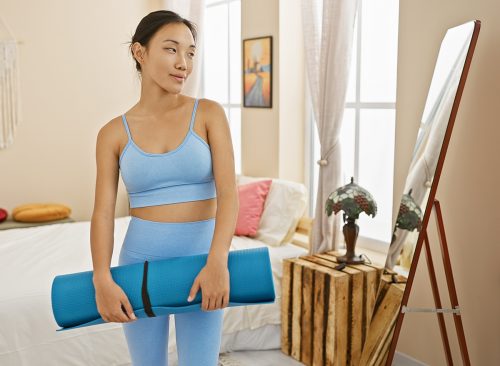 A young asian woman holding a yoga mat, styled in athleisure wear, poised in a tidy, sunlit bedroom interior.