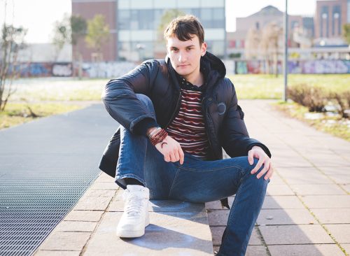young handsome man in the city seated on a sidewalk, wearing blue jeans and a blue jacket.