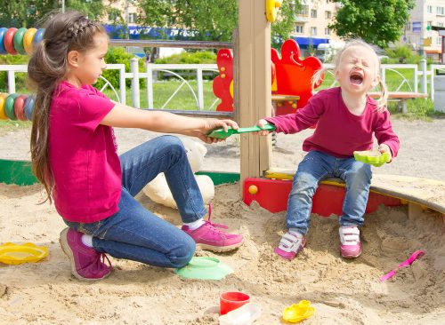 Conflict on the playground. Two sisters fighting over a toy shovel in the sandbox. Kid sister crying all throat