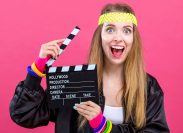 Woman in 1980's fashion holding a film movie slate board clipper on a pink background