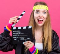 Woman in 1980's fashion holding a film movie slate board clipper on a pink background