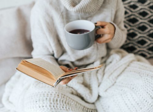Woman hands holding a cup of tea, while she sits in her sofa and reads book