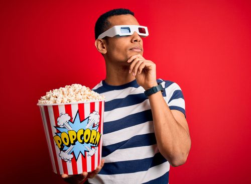 Young handsome african american man watching movie using 3d glasses eating popcorns with hand on chin thinking about question, pensive expression. Smiling with thoughtful face. Doubt concept.