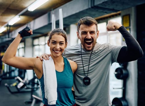 Happy athletic woman and her fitness instructor flexing muscles in a gym while looking at camera.